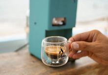 Turquoise coffee pod machine serving a cup of espresso with hand holding the glass cup.